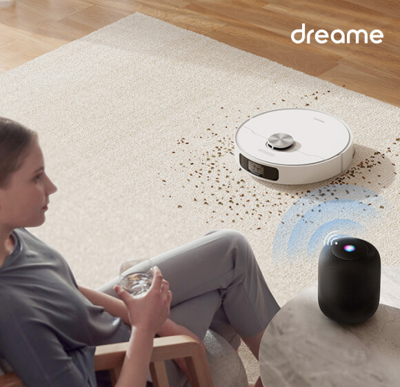 ein Reinigungsroboter saugt Krümel vom Teppich auf, während eine Frau daneben sitzt und Musik aus einem Bluetooth-Lautsprecher hört